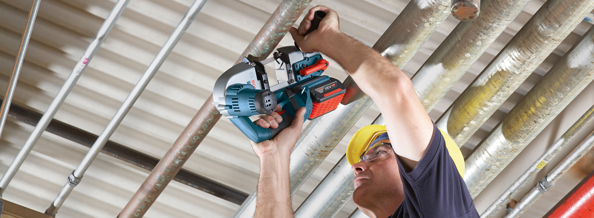 作業員が電動のこぎりを使って天井の金属パイプを切断しています。彼は黄色のヘルメットと保護メガネを着用しています。背景にはテクスチャーのある天井と、さまざまな色とサイズの金属パイプがいくつかあります。