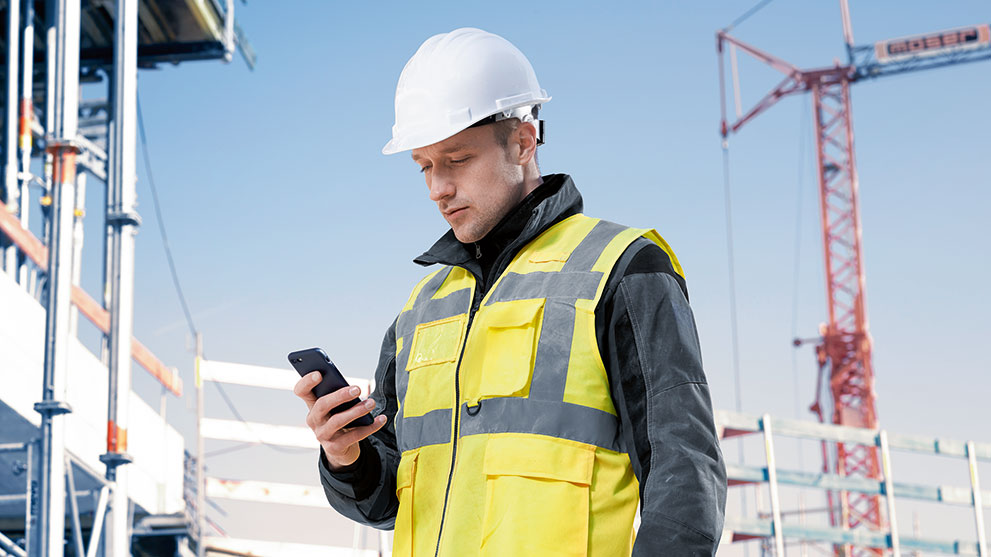 黄色のベストとヘルメットを着た建設作業員がスマートフォンを見ています。