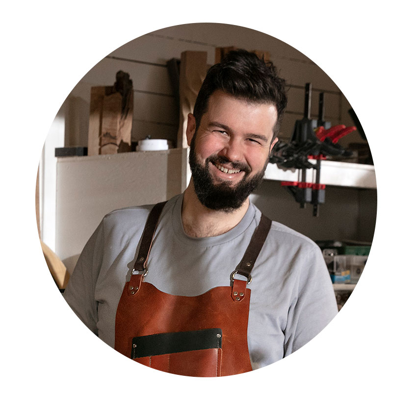 レザーエプロンを着た笑顔の男性が作業場に立っています。