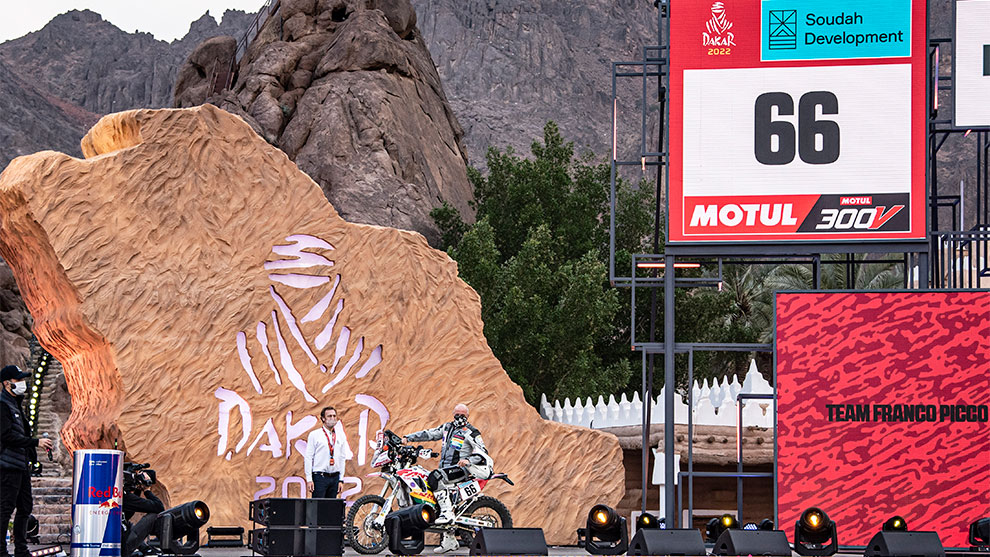 バイクライダーが砂漠の大きなダカールの看板の前に立っています。