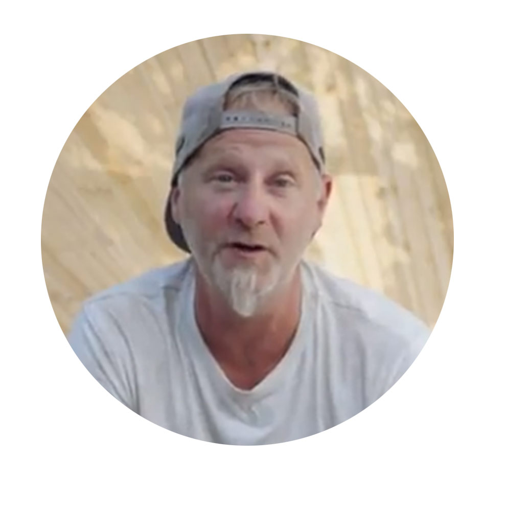 短髪で帽子をかぶった男性が木の構造物の前で微笑んでいます。