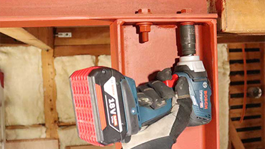 赤いバッテリーを搭載したボッシュプロフェッショナルのコードレスドライバーが金属製の支持具に取り付けられています。背景には木製の壁と断熱材が見えます。